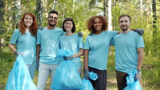 Jovens voluntários alegres com sacos de lixo em pé na floresta abraçando sorrindo
 - Filmagem, Vídeo