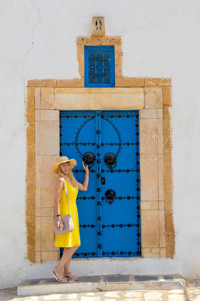 Молодая женщина в желтом платье перед традиционной дверью с узором и плиткой в Сиди-Бу-Саид, Тунис, Тунис
 - Фото, изображение