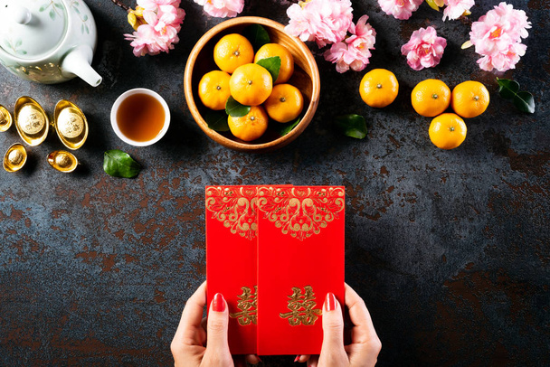 Chinese new year festival decorations. Woman hand holding pow or red packet, orange and gold ingots on a black stone background. Chinese characters FU means fortune good luck, wealth, money flow. - Фото, зображення