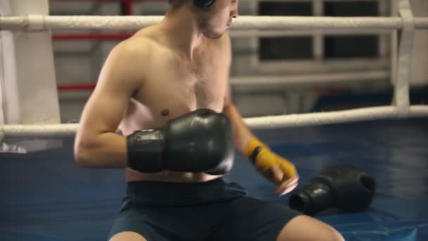 Boxtraining - ein tätowierter Mann sitzt auf dem Ring - setzt Kopfhörer und Handschuhe auf - Filmmaterial, Video