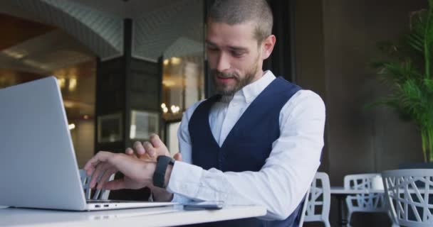 Front view of a young smiling Caucasian businessman in a cafe, checking time on his smartwatch while working on laptop computer, slow motion - Filmati, video