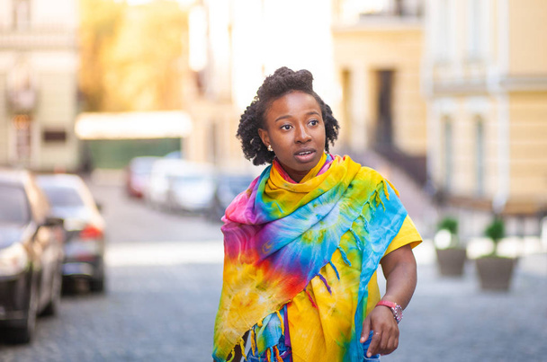 A beautiful African American girl stands on a city street, in a bright yellow shawl. - Фото, зображення