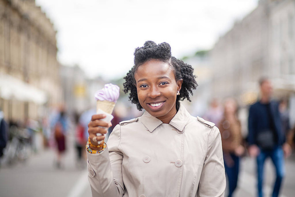 ライフスタイルストリートガール。夏の通りで日焼けしたコートを着てアイスクリームを食べる美しくスタイリッシュな黒の女の子 - 写真・画像