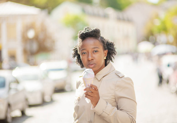 Lifestyle street girl. Beautiful and stylish black girl dressed in a tan coat on a summer street eating ice cream - Фото, зображення