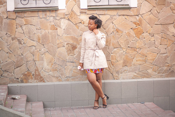 Lifestyle street girl. Beautiful and stylish black girl dressed in a tan coat on a summer street.  - Photo, Image