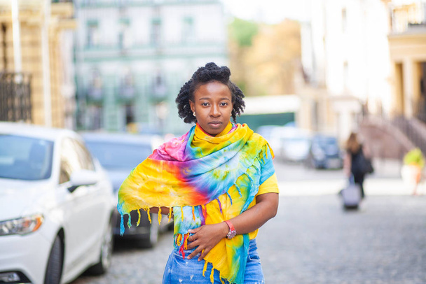 Μια όμορφη Αφρο-Αμερικανίδα στέκεται σε ένα δρόμο της πόλης, με ένα φωτεινό κίτρινο σάλι. - Φωτογραφία, εικόνα