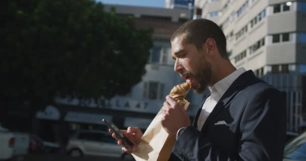 Side view of a young Caucasian businessman on the go in the city, eating and using phone in the street with buildings in the background, slow motion - Felvétel, videó