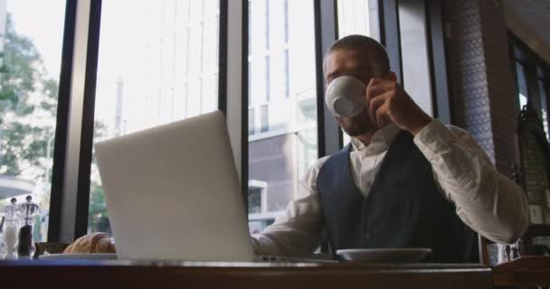 Low angle front view of a young Caucasian businessman in a cafe, using laptop computer and drinking coffee, slow motion - 映像、動画