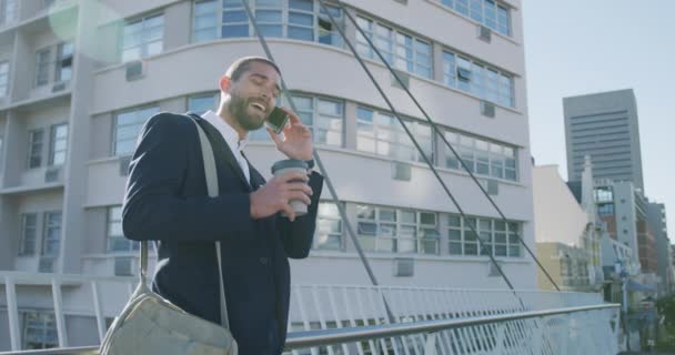 Side view of a young smiling Caucasian businessman on the go in the city, drinking takeaway coffee and talking on the smartphone in the street with buildings in the background, slow motion - Záběry, video