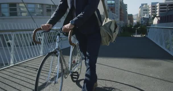 Front view of a young Caucasian businessman on the go in the city, walking with a bike in the street with buildings in the background, slow motion - Footage, Video