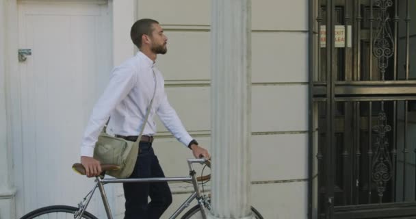 Side view of a young Caucasian businessman on the go in the city, walking with a bike in the street with building in the background, slow motion - Video
