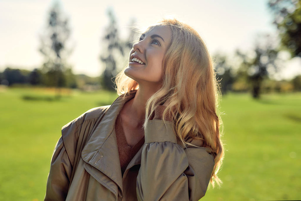 Sunny day. Portrait of young and happy attractive woman in beige cloak smiling and looking up while standing in the middle of the park - Photo, Image