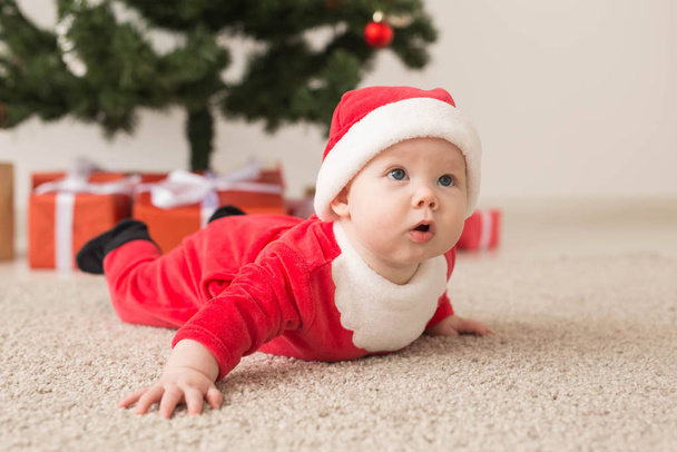 Cute baby girl wearing santa claus suit crawling on floor over Christmas tree. Holiday season. - Photo, Image