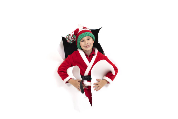 Малыш Санта Клаус выглядывает из дыры в бумаге. Мальчик в новогоднем костюме улыбается. Праздничный. Рождественские праздники
 - Фото, изображение