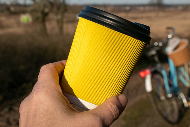 V ruce žlutý papírový zvlněný šálek s kávou na pozadí přírody a na kole - Fotografie, Obrázek
