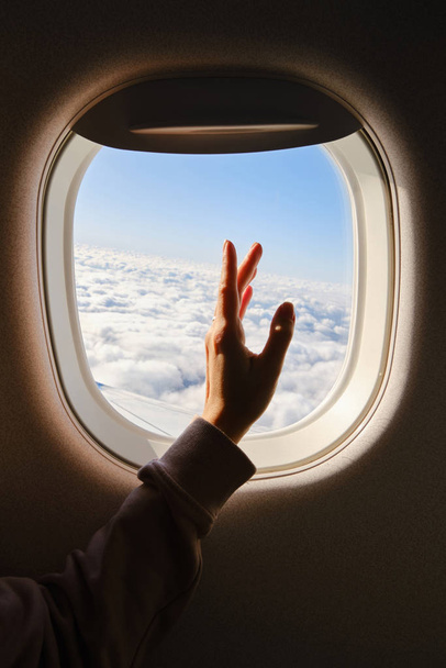 Woman looks out the window of an airplane. Hand near the porthole. Girl on the plane. Airplane wing, scenic view. Beautiful cloud, blue sky. Traveling by plane, adventure. Air transport. Aviation - Foto, afbeelding