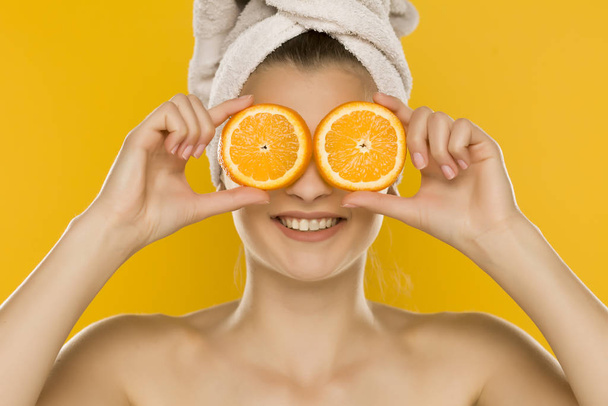 Young beautiful woman with towel on her head holding slices of oranges in front of her eyes on yellow background - Foto, imagen
