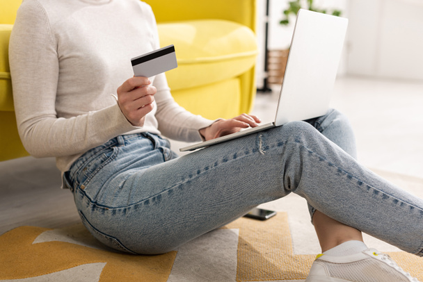 Cropped view of young woman holding credit card and using laptop on floor in living room - Fotografie, Obrázek