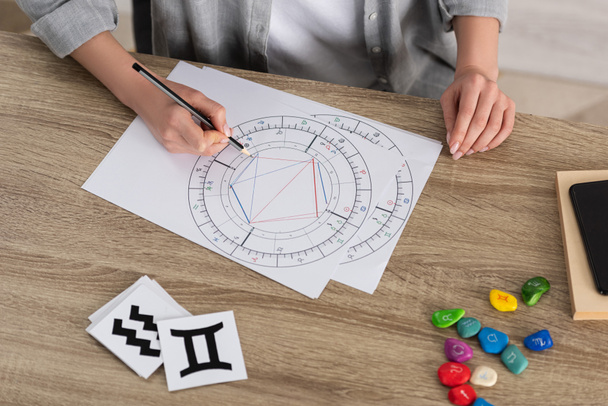 Cropped view of woman drawing natal chart by stones and papers with zodiac signs on table - Photo, Image