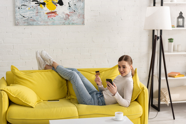 Smiling young woman holding credit card and using smartphone on sofa at home - Фото, изображение