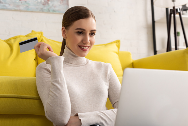 Όμορφο χαμογελαστό κορίτσι κρατώντας πιστωτική κάρτα και κοιτάζοντας φορητό υπολογιστή στο σαλόνι - Φωτογραφία, εικόνα