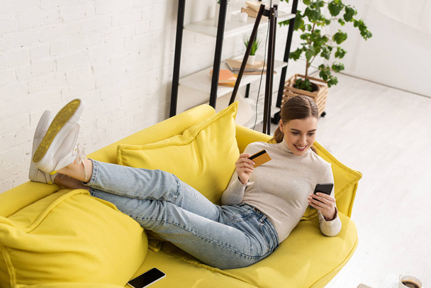Menina sorridente atraente segurando cartão de crédito e usando smartphone no sofá em casa
 - Foto, Imagem