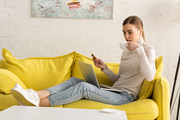 Young woman drinking coffee while holding credit card and using laptop on sofa - Foto, afbeelding