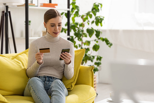 Smiling girl holding credit card and using smartphone on sofa in living room - Photo, Image