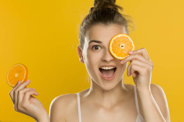 Νεαρή όμορφη γυναίκα κρατώντας φέτα πορτοκαλιού μπροστά από το μάτι της σε κίτρινο φόντο - Φωτογραφία, εικόνα