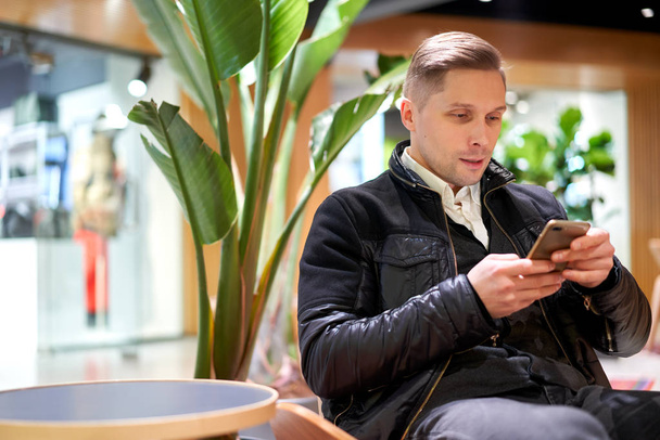 Man in leather jacket with smartphone in his hands sitting on chair in store against green plant - Foto, Bild