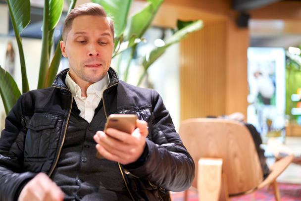 Man in leather jacket with smartphone sitting on chair in store against green plant - Photo, Image