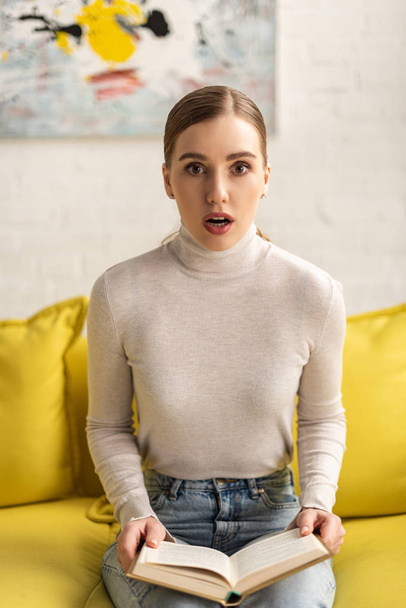 Shocked young woman looking at camera and holding book on sofa - 写真・画像