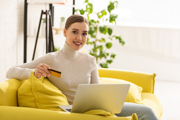 Mladá žena se usmívá do kamery, zatímco drží kreditní kartu a pomocí notebooku na gauči - Fotografie, Obrázek