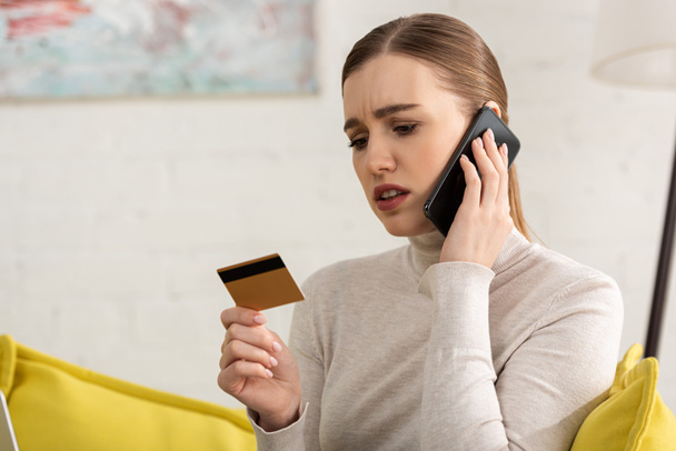 Mujer joven preocupada sosteniendo la tarjeta de crédito y hablando en el teléfono inteligente en casa
 - Foto, Imagen