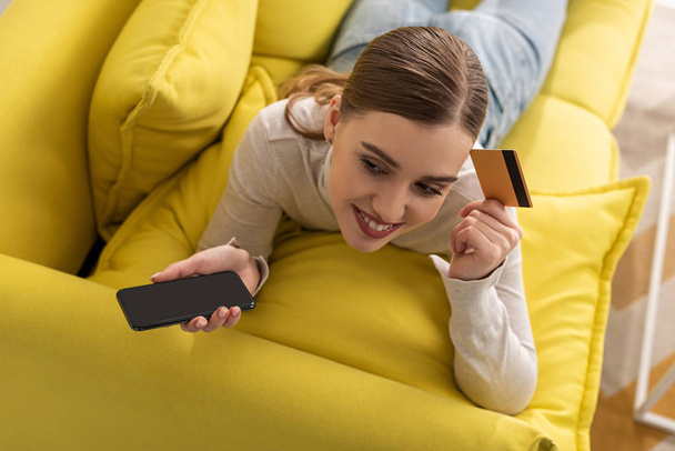Вид сверху на улыбающуюся женщину, держащую кредитку и смартфон, лежащую на диване
 - Фото, изображение