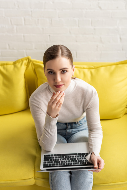 Pensive woman looking at camera while using laptop on couch  - Photo, Image