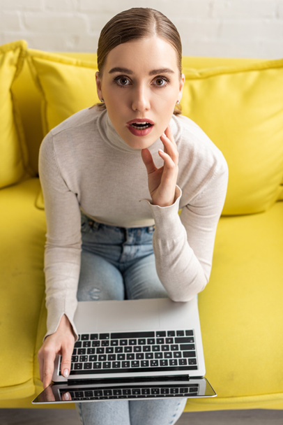 High angle view of shocked woman with laptop looking at camera on sofa - Foto, afbeelding