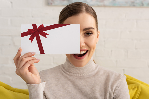 Attractive exited girl covering face with gift card and looking at camera - Fotografie, Obrázek