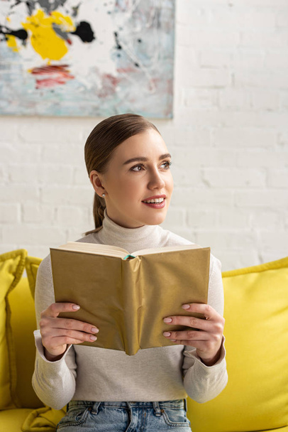 Smiling woman holding book and looking away on couch in living room - Foto, Bild
