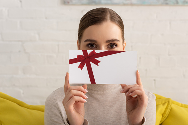 Young woman obscured face with gift card and looking at camera on sofa - Photo, Image