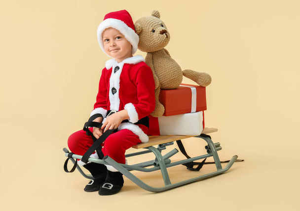 Cute little boy in Santa costume, with sledges and Christmas gifts on color background - Фото, изображение