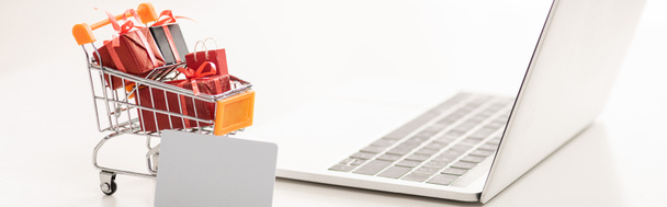 Lelukärry lahjapakkauksissa kannettavan tietokoneen ja luottokortin vieressä valkoisella pinnalla, panoraama laukaus
 - Valokuva, kuva