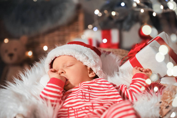 Cute little baby with Santa Claus hat and Christmas gift sleeping in basket at home - Foto, imagen