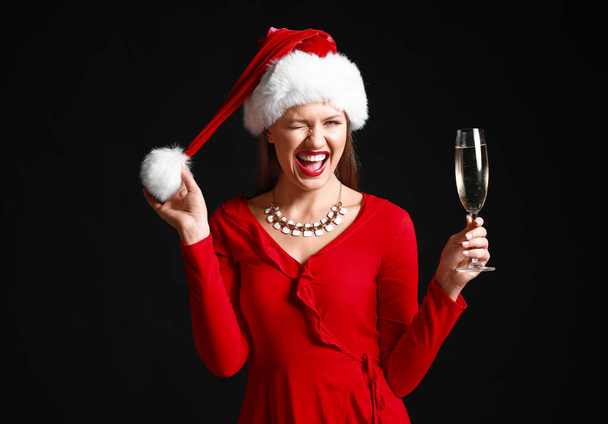 Happy young woman in Santa Claus hat and with glass of champagne on dark background - 写真・画像