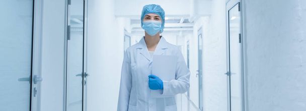 Γυναίκα σε προστατευτική ιατρική φθορά διέρχεται από άδειο εργαστηριακό διάδρομο - Φωτογραφία, εικόνα