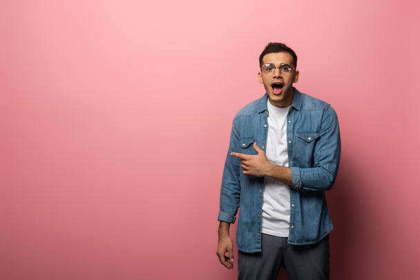 Young shocked man pointing with finger and looking at camera on pink background - Fotoğraf, Görsel