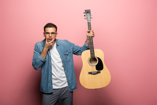 Handsome man grimacing at camera and holding acoustic guitar on pink background - Foto, Imagen