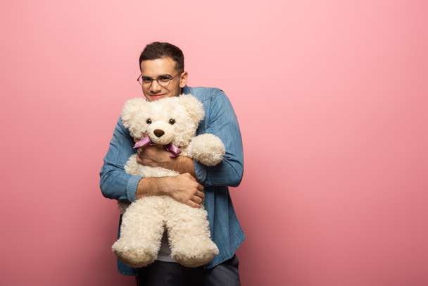 Όμορφος χαμογελαστός άντρας αγκαλιάζει αρκουδάκι και κοιτάζει την κάμερα σε ροζ φόντο - Φωτογραφία, εικόνα