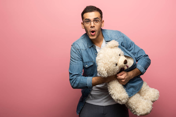 Shocked man looking at camera while holding teddy bear on pink background - Фото, изображение