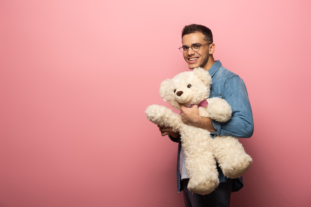 Young man smiling at camera and holding teddy bear on pink background - Фото, изображение